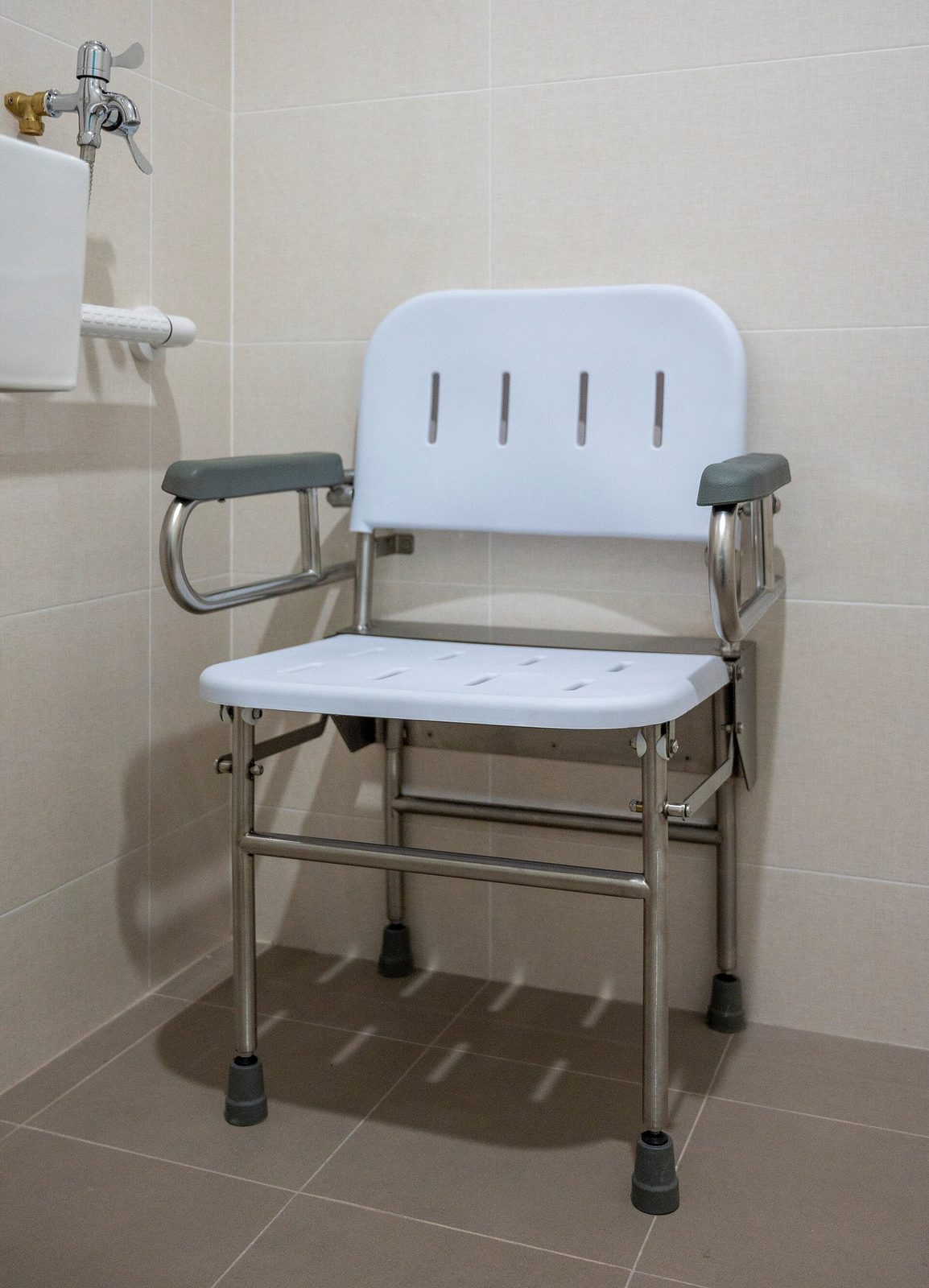 Opklapbare douchestoel met armleuningen tegen een getegelde wand, met een witte steunbeugel ernaast