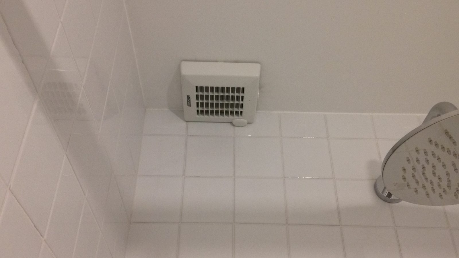 Witte afzuigventilator boven de wandtegels in een douche, met een ronde regendouchekop op de voorgrond